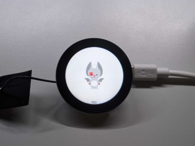 Round display with a black bezel around a white image with the Golioth Echo mascot at the center. A USB cable is plugged into the device on the right side of the screen.